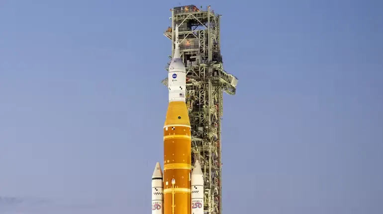 NASA Artemis rocket with orange and white sections on launch pad against clear blue sky, tall service tower visible